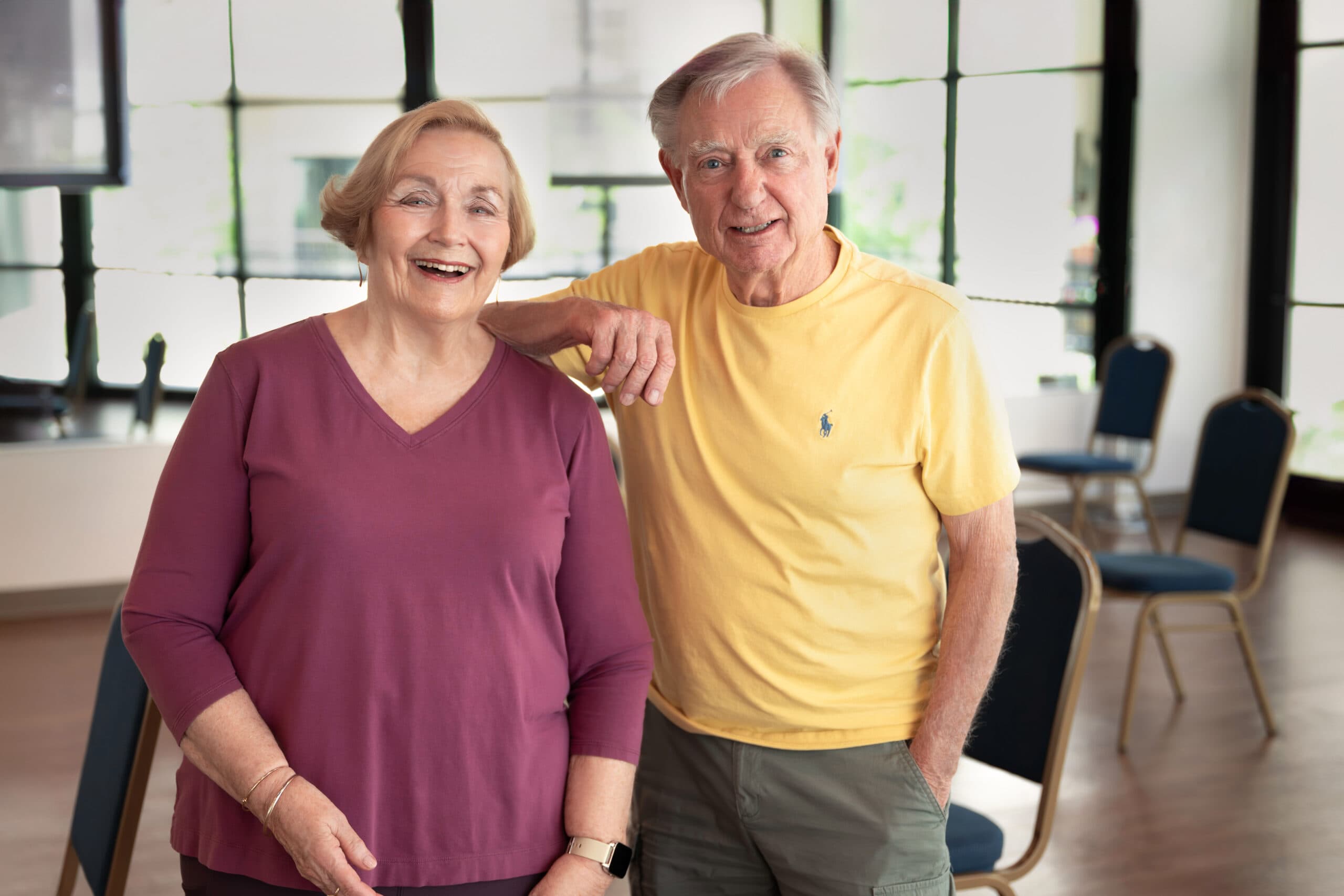 A smiling older couple standing arm in arm in a bright activity room with chairs arranged behind them.