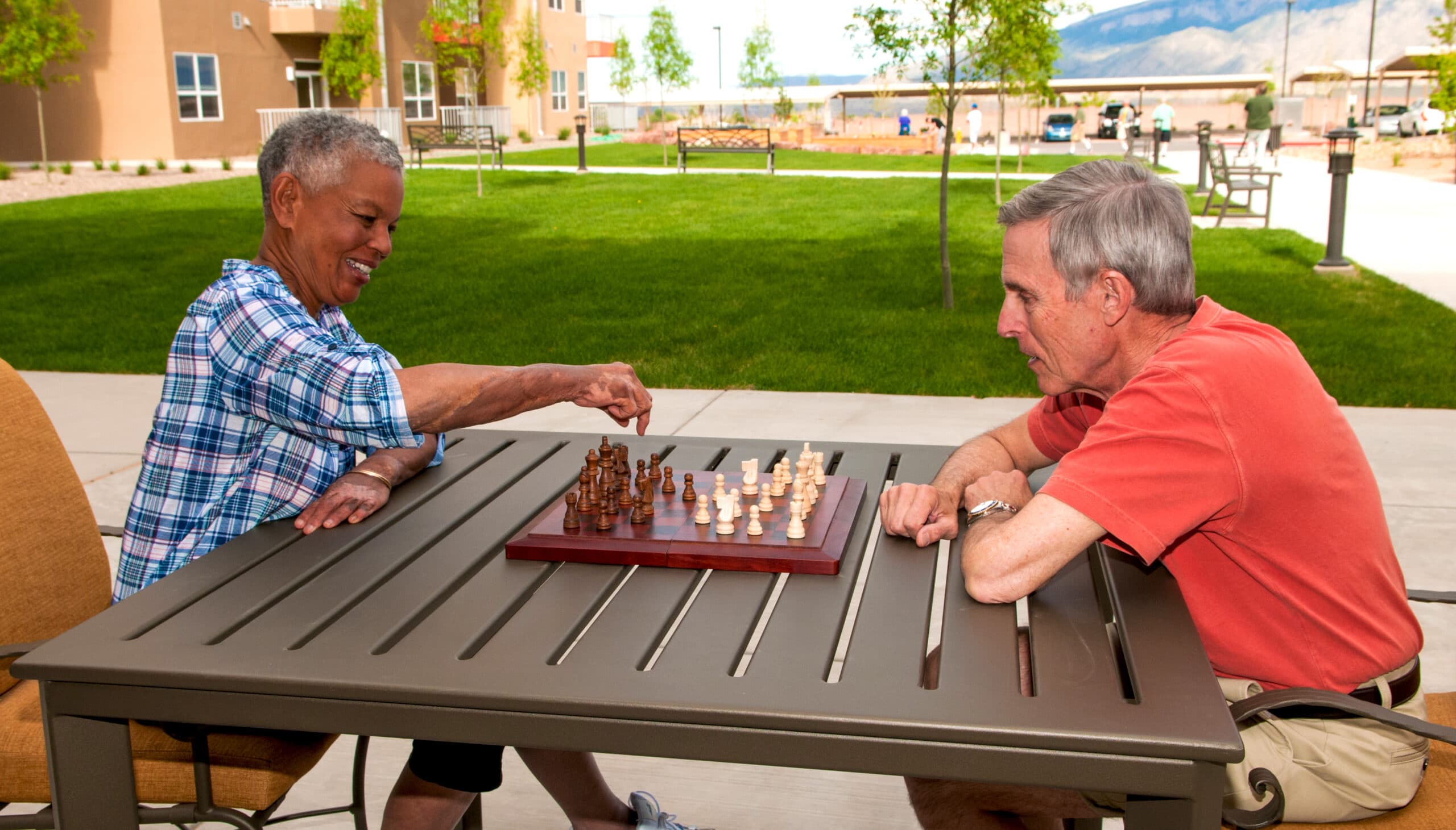 Two seniors seated at an outdoor table under trees playing chess on a wooden chessboard in a courtyard.