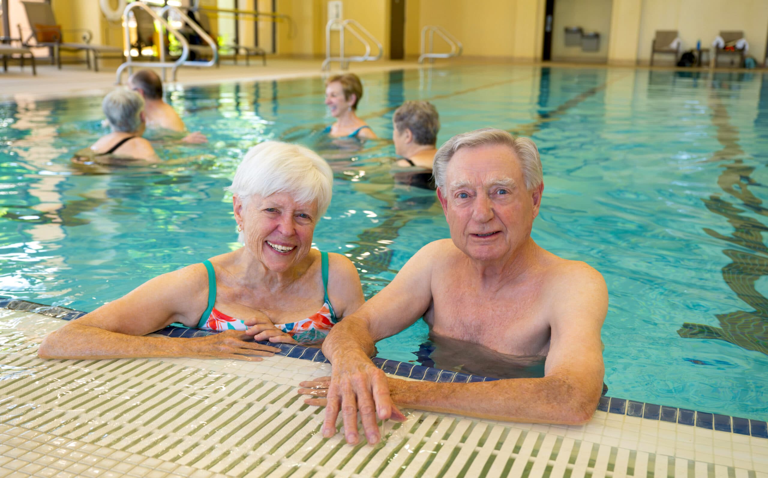 An older man and woman standing chest-deep in an indoor pool smiling at the camera while others exercise in the background.