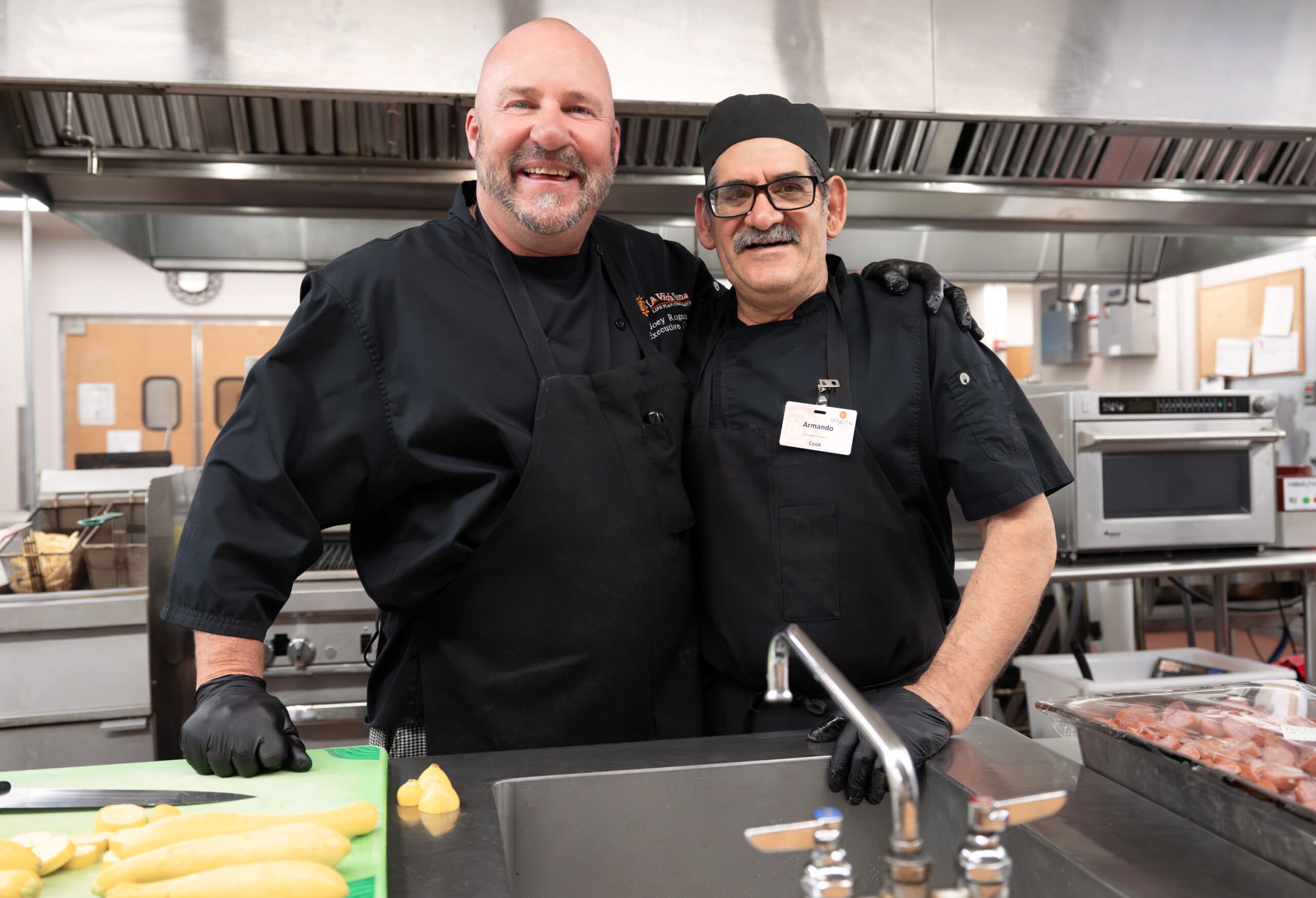 Two commercial chefs in black uniforms and gloves standing side by side in a stainless-steel kitchen, smiling at the camera.