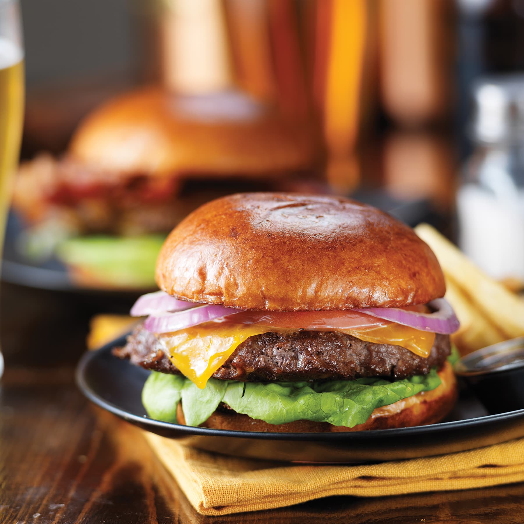 A juicy cheeseburger on a plate, with melted cheddar, red onion, lettuce and tomato on a brioche bun, and fries blurred in the background.