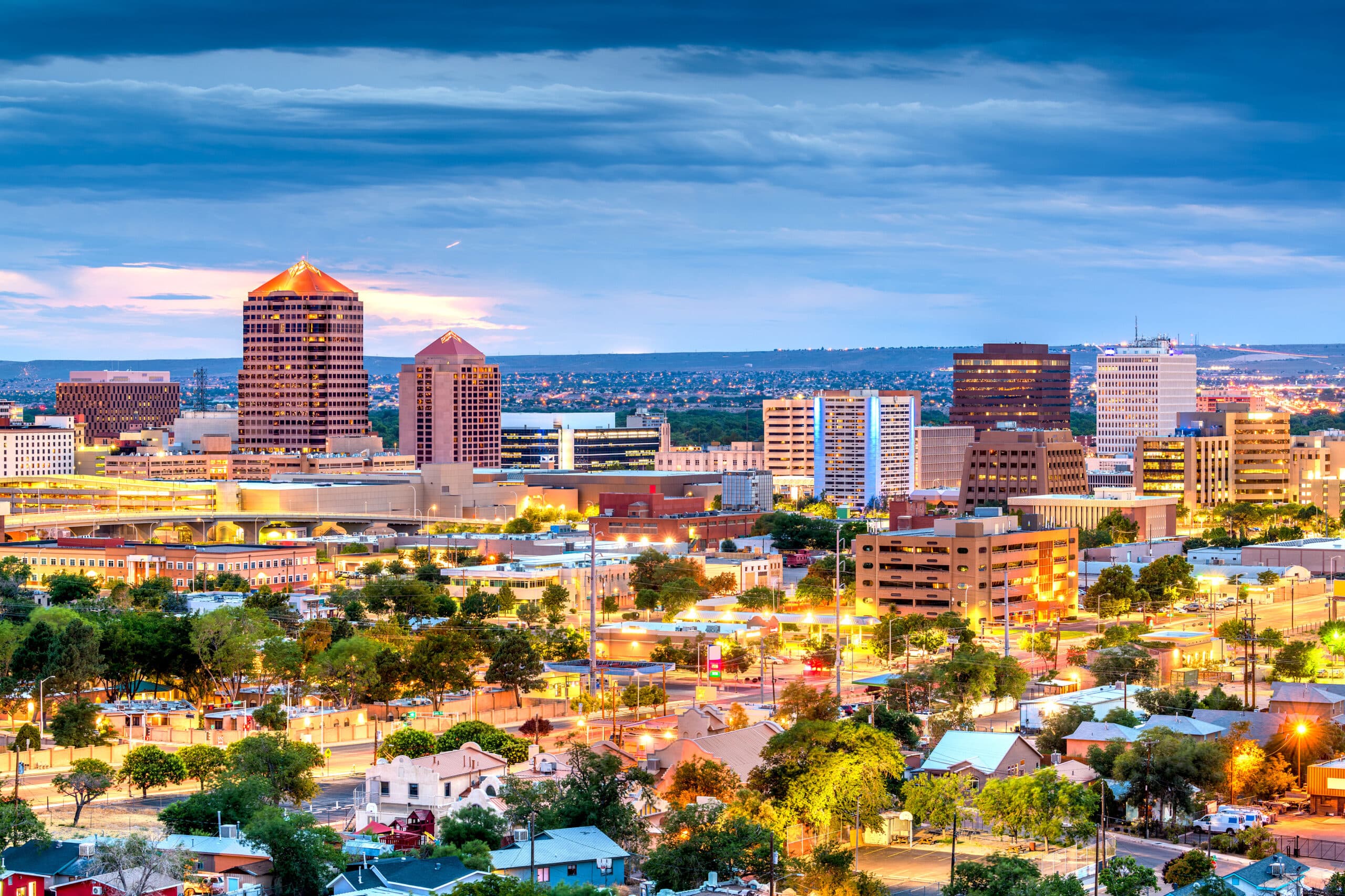 Twilight view of downtown Albuquerque, with illuminated modern high-rises against the distant Sandia Mountains under a deepening blue sky.