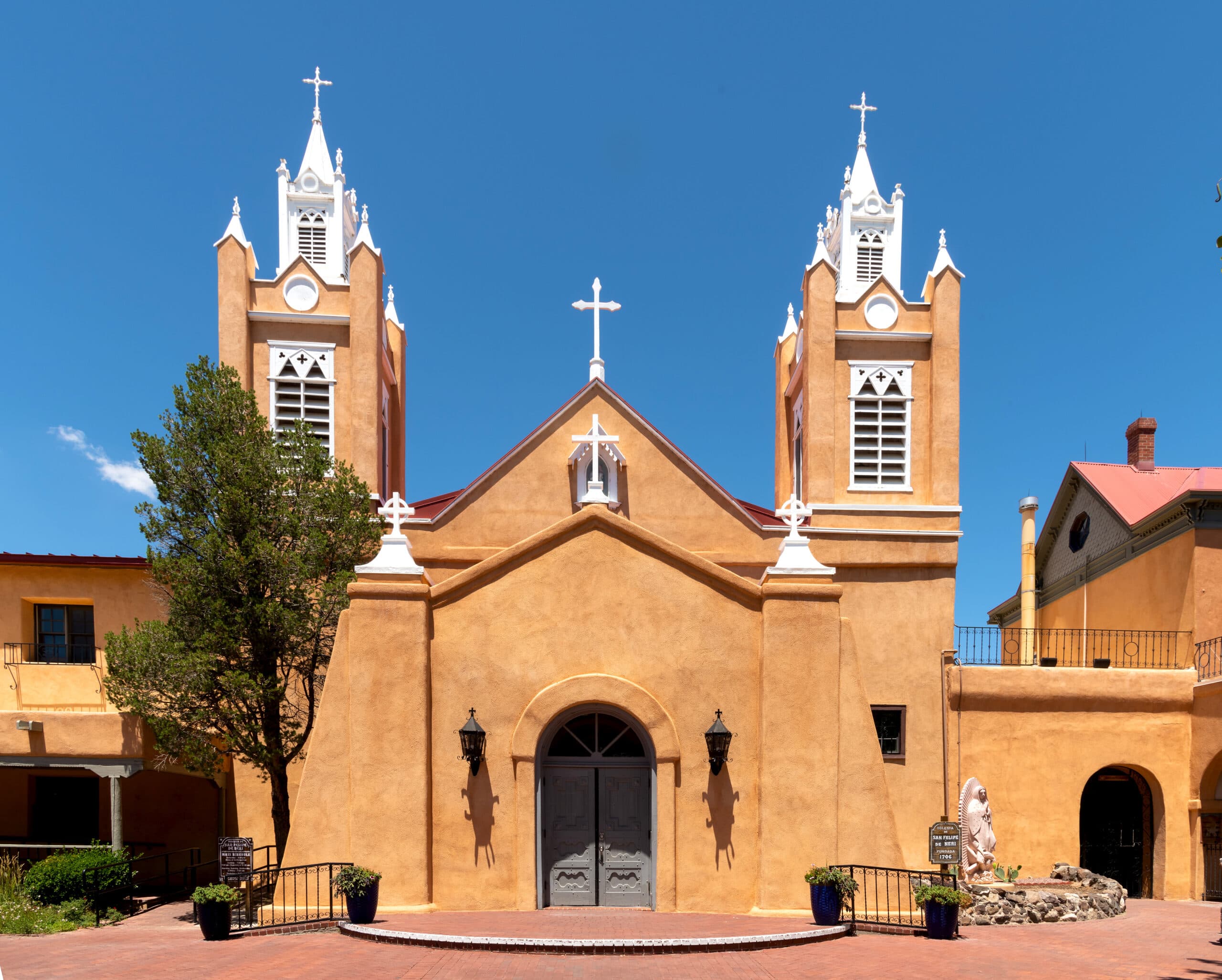 Front facade of the adobe San Felipe de Neri Church in Old Town Albuquerque, featuring two white bell towers and a central cross against a clear blue sky.