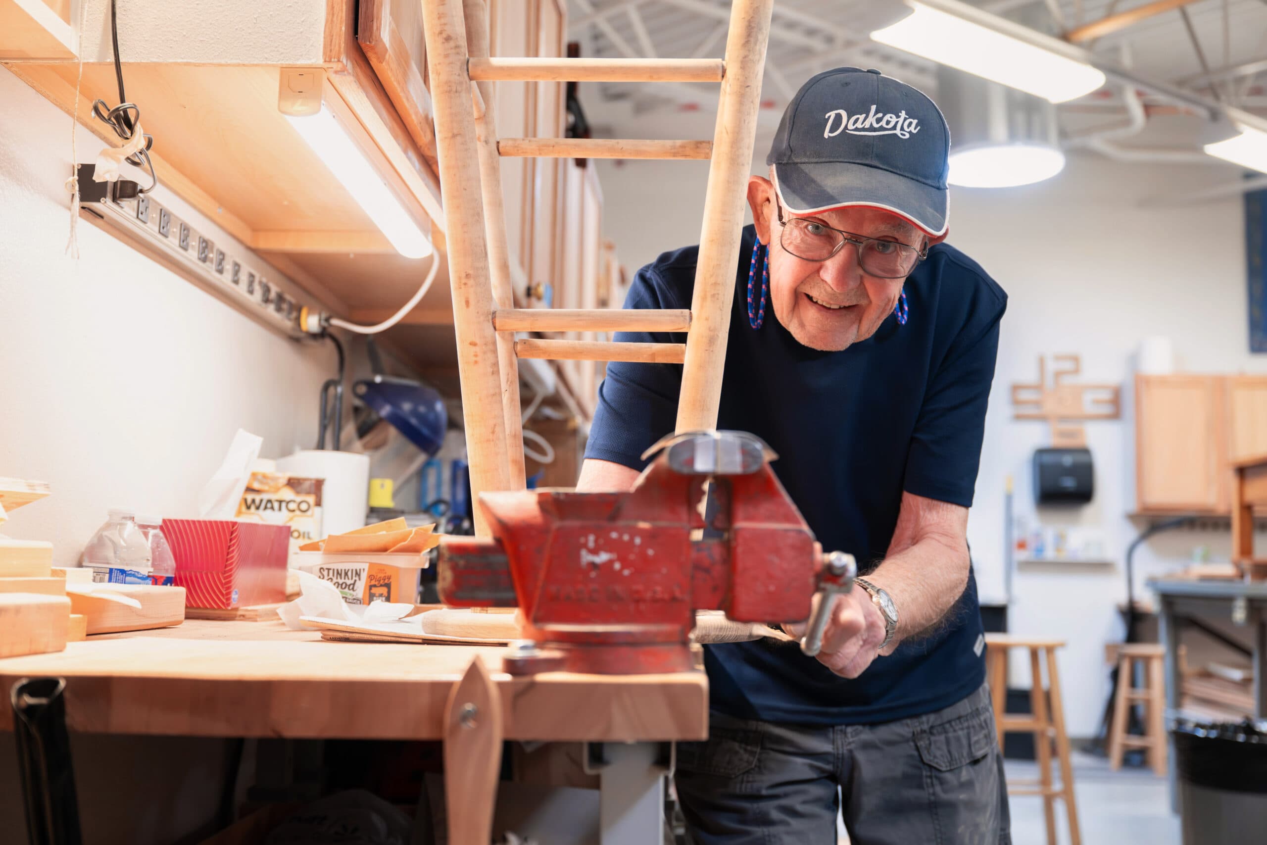 A senior man in glasses standing beside a wooden cabinet in a workshop, smiling with a pegboard of woodworking tools in the background.