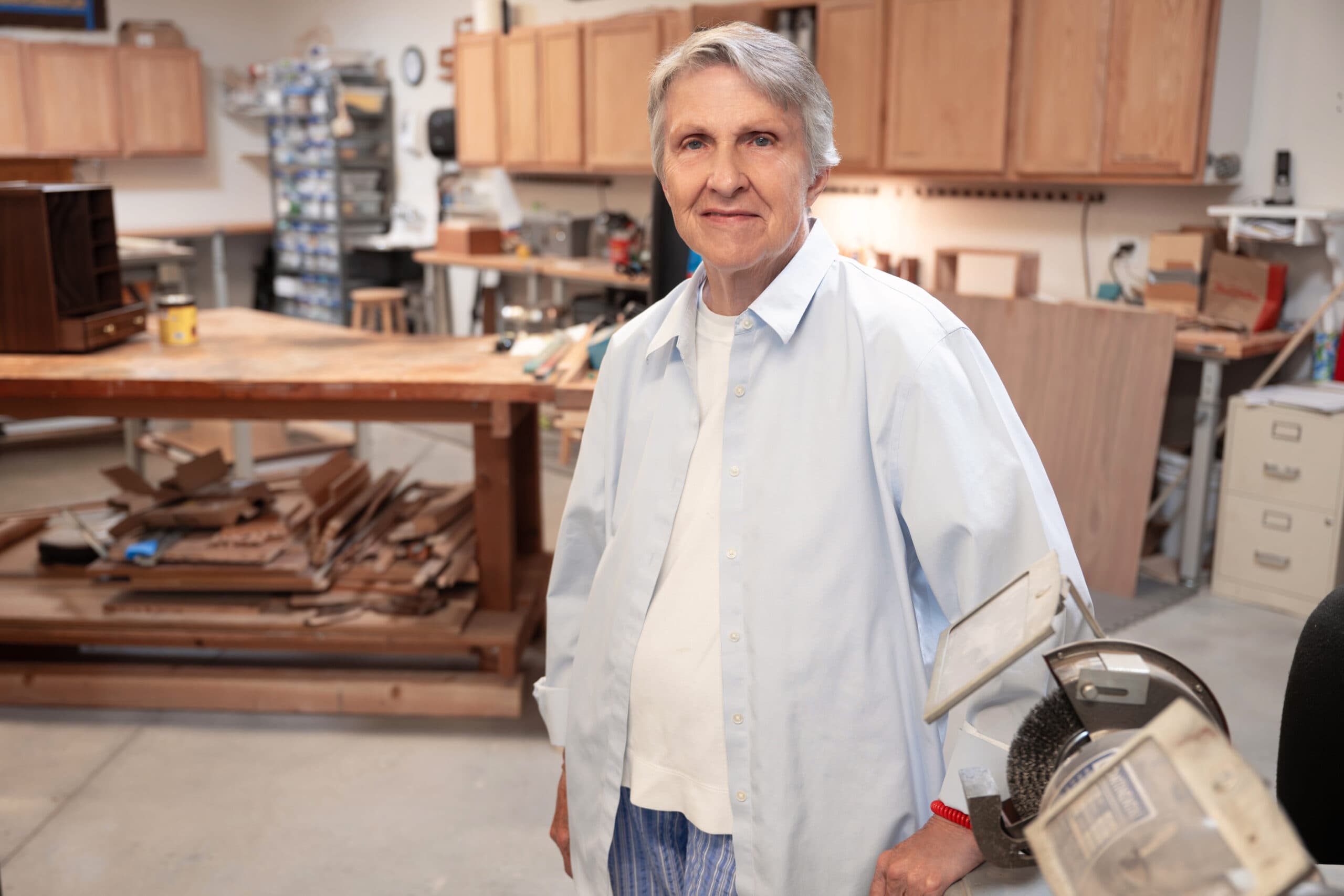 An elderly woman standing in a wood shop, posing with woodworking machinery and tools visible around them.