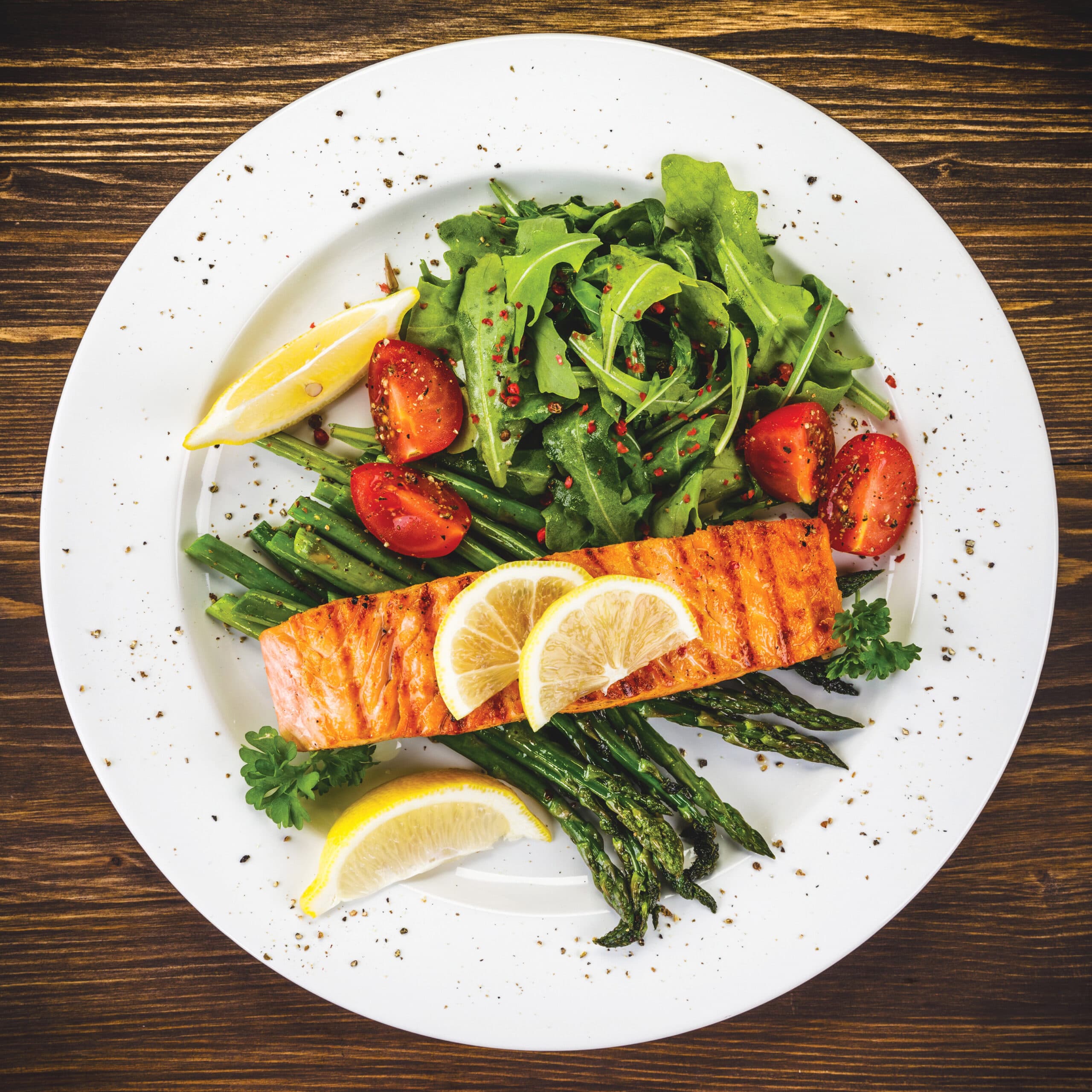 Top-down shot of a salmon fillet on a bed of asparagus, garnished with arugula, cherry tomatoes and lemon wedges on a white plate.